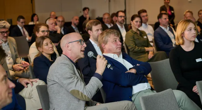 Ein Saal voll sitzender Menschen, im Vordergrund ein Herr im grauen Anzug der sich mit einem Mikrofon zu Wort meldet.