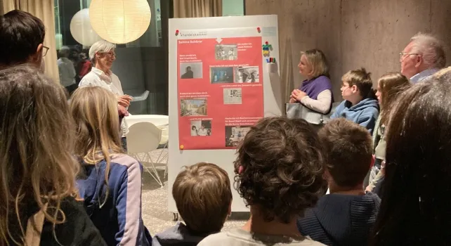 Das Bild zeigt Eltern und Kinder, die einem Mann vor seinem Plakatvortrag zuhören