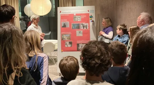 Das Bild zeigt Eltern und Kinder, die einem Mann vor seinem Plakatvortrag zuhören